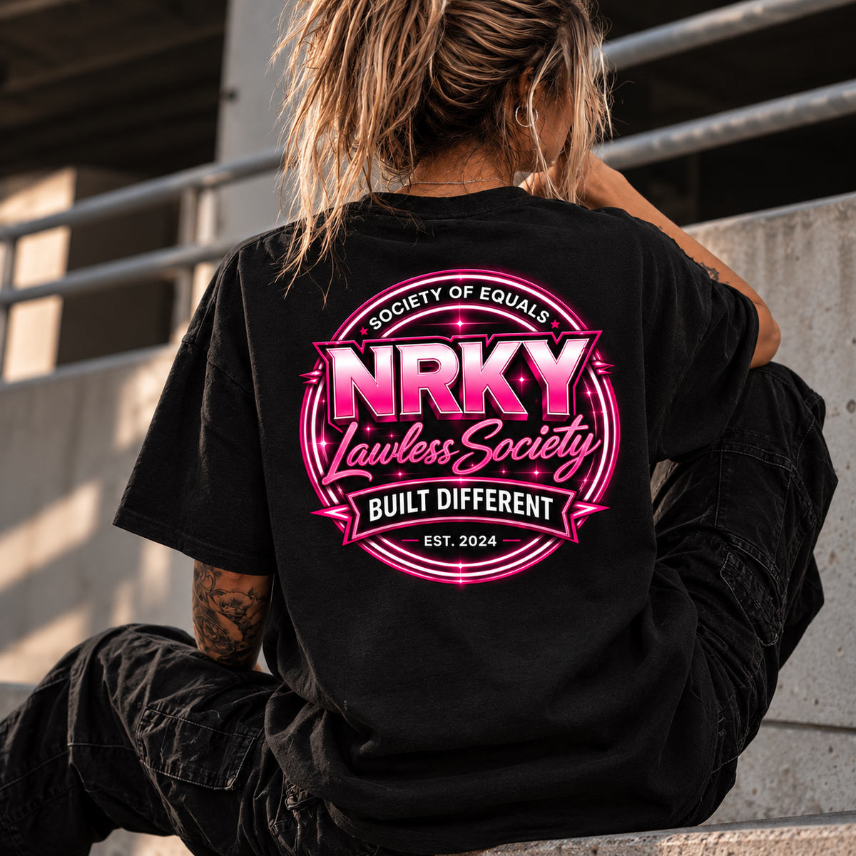 Women's Sinister Pink tee with NRKY Lawless Society hot pink back print, seated lifestyle photo